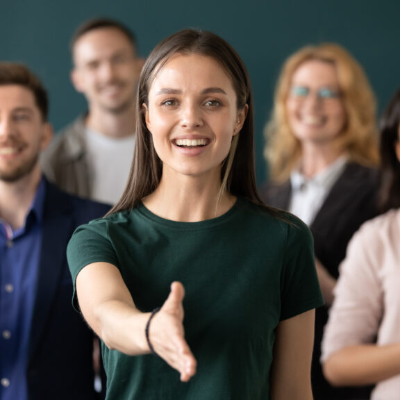 Friendly young company woman representative holds out her hand for handshake welcoming customer smiling looking at camera posing together with diverse colleagues, sales manager greeting client concept