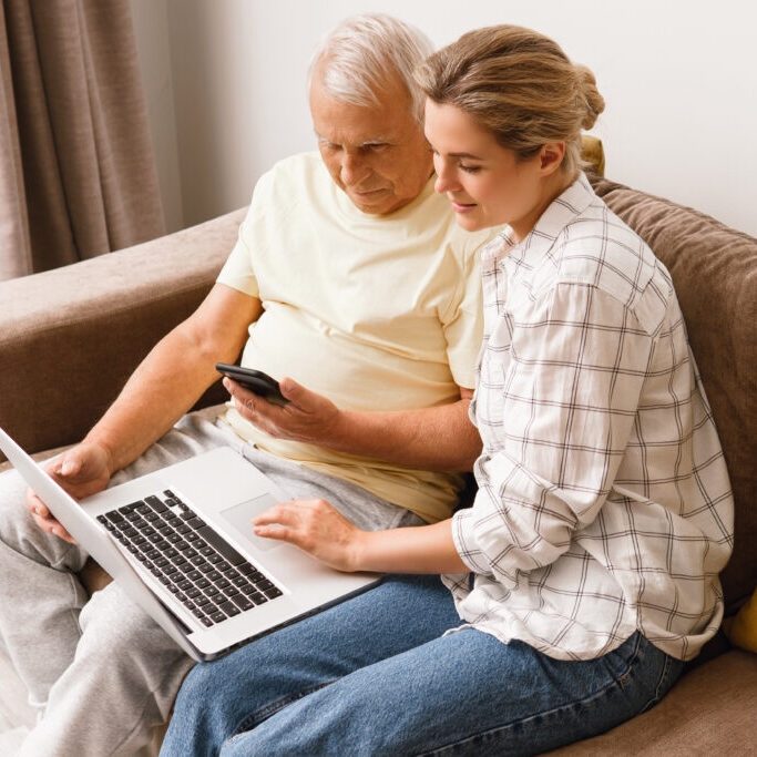 Young woman explaining to elderly man how to use laptop and smatphone